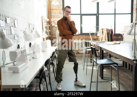 Jeune designer sérieux de mode ou tailleur avec prothèse au lieu de une jambe se tenant près du lieu de travail devant l'appareil photo à l'intérieur grand atelier Banque D'Images