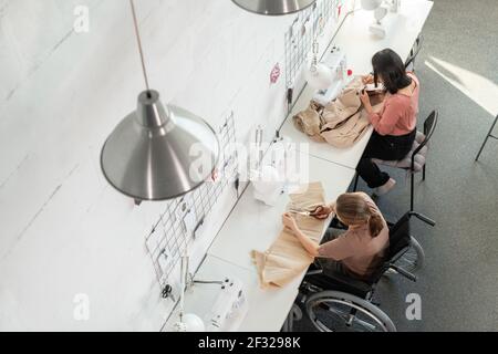 Une jeune couturière et sa collègue de sexe féminin et inadapté assis sur le lieu de travail intérieur grand atelier contemporain et vêtements de couture Banque D'Images