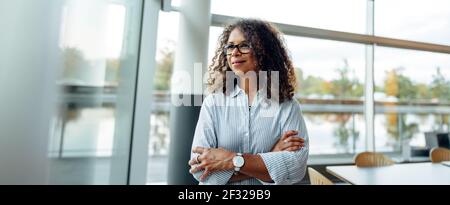 Femme entrepreneur debout à côté d'une fenêtre dans le bureau. Femme mûre debout au bureau avec les bras croisés regardant à l'extérieur de la fenêtre. Banque D'Images