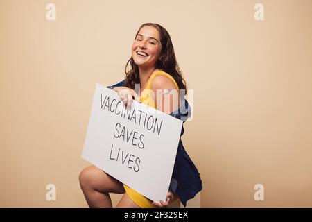 Jolie femelle après avoir reçu un vaccin sur le bras assis sur fond marron. Femme tenant une bannière « la vaccination sauve des vies » regardant la caméra et Banque D'Images