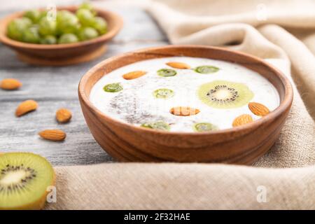 Yaourt au kiwi, à la groseille, au chia et aux amandes dans un bol en bois sur fond en bois gris et linge de maison. Vue latérale, gros plan, mise au point sélective. Banque D'Images