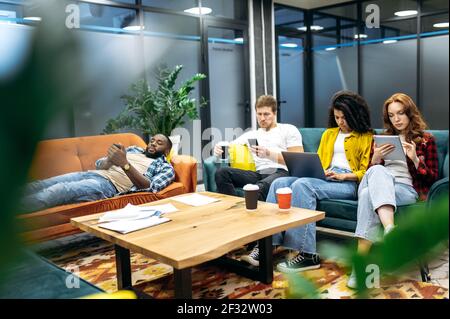 Jeunes gens d'affaires multiraciaux adultes en pause dans un bureau moderne. Collègues ou étudiants élégants assis sur le canapé, utilisant des gadgets, naviguer sur Internet, se reposer après un travail ou un travail Banque D'Images