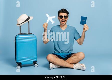 Excitée joyeuse caucasien touriste mâle dans des lunettes de soleil se trouve sur fond bleu isolé, tenant passeport d'une main, l'avion dans l'autre, la valise bleue est debout à proximité, vacances et concept de voyage Banque D'Images