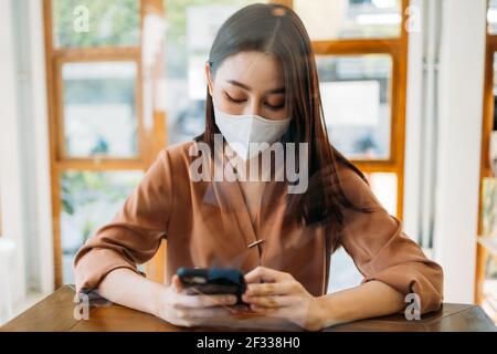 Belle jeune femme asiatique assise dans un café moderne portant un covid-19 masque de protection du visage utilisation et navigation dans le smartphone tout en vous relaxant Banque D'Images