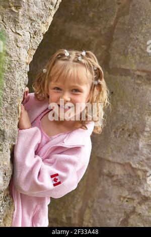 Bonne fille blonde souriant et regardant l'appareil-photo tout en regardant de derrière le vieux mur de pierre, pendant une visite de Burgruine Leofels, Ilshofen Banque D'Images