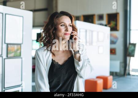 Charmante jeune femme d'affaires artiste designer en costume blanc utilisant téléphone mobile dans la galerie d'art moderne Banque D'Images