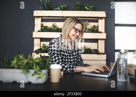 jolie jeune femme d'affaires avec une chemise à carreaux noir et blanc et des lunettes noires est assise dans un bureau écologique et durable et travaille sur un ordinateur portable Banque D'Images