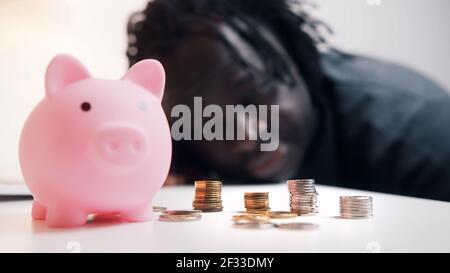 Décision financière, investissements et économies. Homme afro-américain regardant dans les pièces de monnaie devant la banque de piggy. Photo de haute qualité Banque D'Images