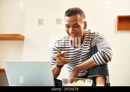Souriant, jeune vietnamien, mangeant des chips et regardant la comédie film sur écran d'ordinateur portable Banque D'Images