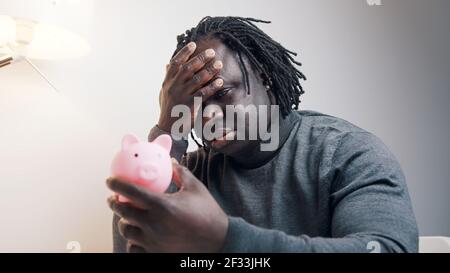 Concept de faillite. Triste homme noir afro-américain regardant la banque de porc vide et secouant sa tête. Photo de haute qualité Banque D'Images
