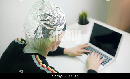 Femme craintive avec un chapeau en aluminium travaillant sur un ordinateur portable. Protection contre les 5 g et la lecture et le contrôle de l'esprit. Photo de haute qualité Banque D'Images