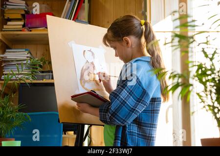 Une jeune fille de dix ans tire avec des peintures sur un chevalet, tenant une tablette informatique dans ses mains Banque D'Images