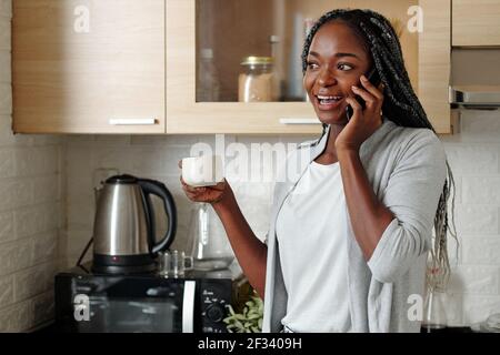 Portrait d'une jeune femme noire heureuse et excitée qui boit le café du matin dans la cuisine et parler au téléphone avec un ami Banque D'Images