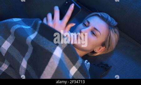 Portrait d'une jeune femme parcourant les médias sociaux tout en se relaxant sur le canapé recouvert d'une couverture. Photo de haute qualité Banque D'Images