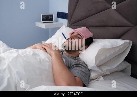 Jeune latino malade au lit avec un thermomètre dans la bouche et un tissu sur la tête. Horloge numérique à l'arrière-plan. Banque D'Images