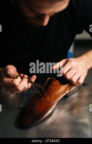 Vue en gros plan du cordonnier mâle barbu qui noue les lacets sur les chaussures en cuir brun clair poli et réparé. Banque D'Images