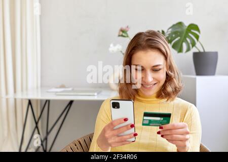 Elle a été ravie d'acheter une carte en plastique et un smartphone chez elle. Femme souriante payant par carte pour un achat en ligne par téléphone Banque D'Images