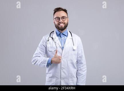 Un médecin de race blanche positif dans une blouse de laboratoire montrant le pouce vers le haut geste sur fond gris de studio Banque D'Images