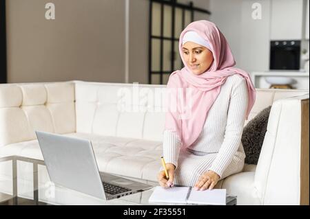 Portrait d'une jeune femme musulmane dans le hijab travaillant à la maison à l'aide d'un ordinateur portable tout en étant assis sur un canapé, écrire des notes, écrire une liste de tâches, travailler à distance, étudier en ligne, indépendant écrivant des détails Banque D'Images