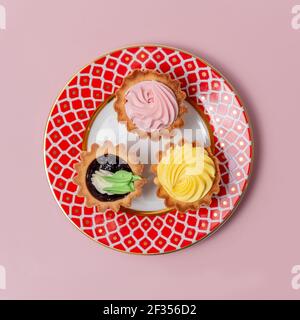 Délicieux petits gâteaux sur une assiette à motif rouge. Trois petits gâteaux différents à la crème rose et jaune Banque D'Images