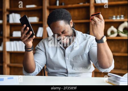 Joyeux homme d'affaires afro-américain assis à un bureau tenant une carte de crédit et un téléphone mobile pour effectuer des transactions en ligne, achetant des articles souhaitée, criant oui, shopping à la maison concept Banque D'Images