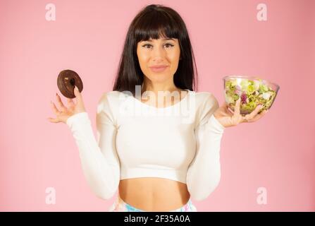 Jeune belle femme brune portant des vêtements de sport tenant des beignes et de la salade isolée sur fond rose. Banque D'Images