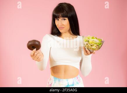 Jeune belle femme brune portant des vêtements de sport tenant des beignes et de la salade isolée sur fond rose. Banque D'Images