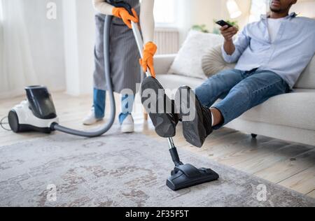 Femme noire passant l'aspirateur sur le sol pendant que son mari regarde la télévision, ne l'aidant pas avec les tâches ménagères, vue rognée Banque D'Images