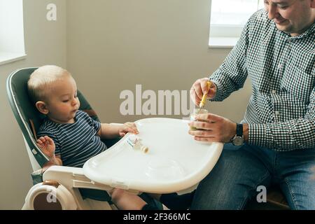 Père caucasien nourrissant un petit garçon mignon avec une purée à la cuillère à la maison, assis à la chaise haute de l'enfant. Famille, nourriture, manger, style de vie, conce authentique Banque D'Images