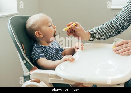 Père caucasien nourrissant un petit garçon mignon avec une purée à la cuillère à la maison, assis à la chaise haute de l'enfant. Famille, nourriture, manger, style de vie, conce authentique Banque D'Images