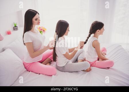 Photo de doux attrayant trois vêtements de nuit de frères et sœurs assis tresse de lit se brosser les cheveux à l'intérieur de la maison Banque D'Images