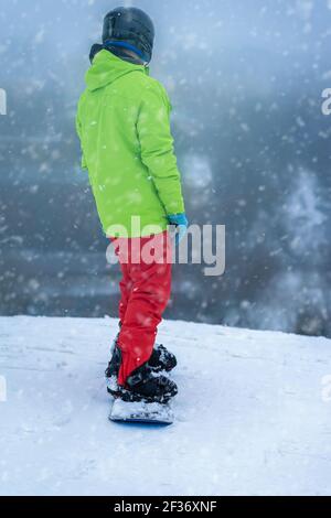 Snowboardeur en blouson vert sportif brillant debout sur son plateau en haut de la pente. Mode de vie actif, sport extrême d'hiver et concept de loisirs Banque D'Images