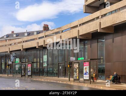 Les abris de bus le long d'une rue juste après une douche de pluie. Un bâtiment moderne en verre se courbe avec la rue et les gens attendent un bus. Banque D'Images