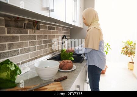 Arabe musulmane en hijab beige debout dans la cuisine et laver les feuilles de salade dans la cuisine avant de préparer le vegan salade par beau temps ensoleillé Banque D'Images