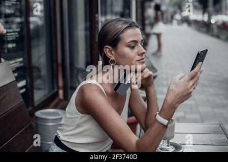 Jeune femme souriante magasiner en ligne en utilisant le téléphone à l'extérieur dans le café. Un beau modèle regarde le téléphone et boit le café en été. Banque D'Images