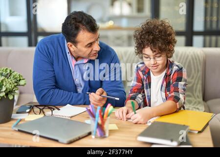 Offrez à votre enfant le meilleur. Un père latin d'âge moyen attentionné vérifie ses devoirs, aide son fils et son enfant à l'école à étudier tout en étant assis au Banque D'Images