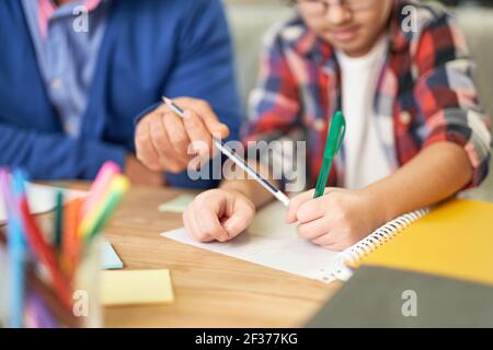 Gros plan des mains d'un père latin d'âge moyen aidant son fils, enfant d'âge scolaire à faire ses devoirs tout en étant assis ensemble au bureau à la maison Banque D'Images