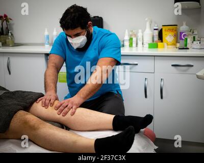 Un physiothérapeute dans un masque et des vêtements bleus traitant un genou du patient Banque D'Images