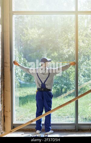 Prise de vue en longueur d'un jeune ouvrier portant une combinaison bleue avec mesurer le ruban pendant les travaux sur le chantier de construction de chalets Banque D'Images
