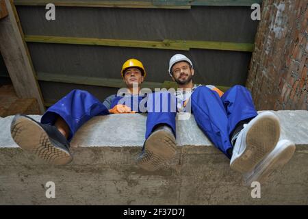 Deux jeunes bâtisseurs attrayants en combinaison bleue et en casque de sécurité regardant vers le bas la caméra, assis sur le sol en béton, se reposant pendant le travail à la maison Banque D'Images