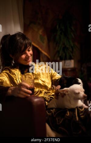 une femme rit aux petits chiens sur les genoux avec un verre à l'intérieur la main à la maison pour se détendre Banque D'Images