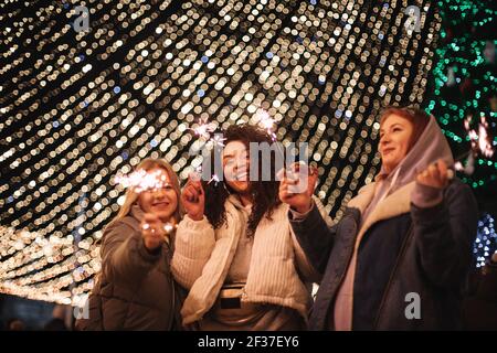 Amies féminines heureuses tenant des étincelles debout près des lumières de noël Banque D'Images