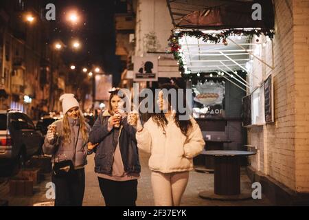 Des amies qui ont des boissons chaudes à marcher dans la rue en ville la nuit Banque D'Images