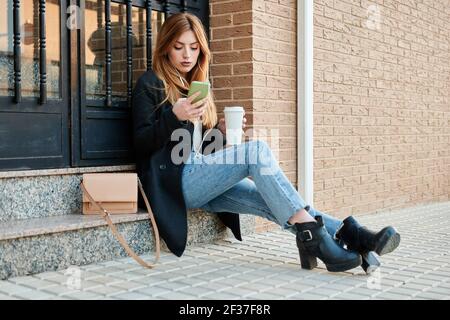Une femme d'affaires blonde boit du café et regarde son téléphone Banque D'Images