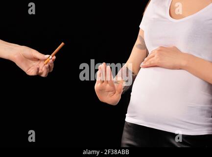 La jeune femme enceinte refuse de prendre la cigarette suggérée sur fond sombre. Concept de mode de vie sain Banque D'Images