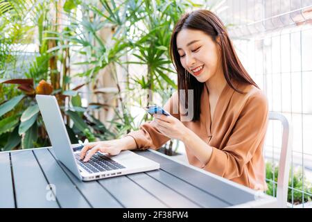 Belle jeune femme asiatique souriant et tenant une carte de crédit bancaire pour le paiement sur ordinateur portable pour les achats en ligne assis dans moderne café Banque D'Images