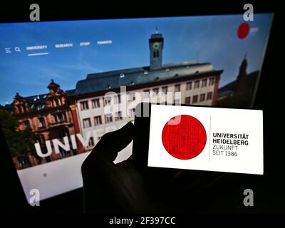 Personne tenant un téléphone portable avec le logo de l'université allemande Ruprecht-Karls-Universität Heidelberg à l'écran en face de la page web. Mise au point sur l'affichage du téléphone. Banque D'Images