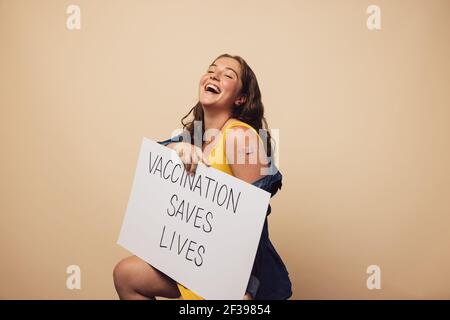 Une femme en train de rire avec une bannière montrant « la vaccination sauve des vies ». Femelle après avoir reçu la vaccination, portant une bannière sur fond marron. Banque D'Images