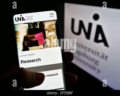 Personne tenant un téléphone portable avec la page web de l'université allemande Universität Augsburg à l'écran devant le logo. Concentrez-vous sur le centre de l'écran du téléphone. Banque D'Images