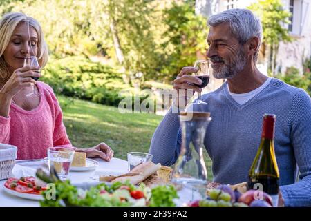 Couple mûr souriant qui sent le vin rouge tout en étant assis à table Banque D'Images